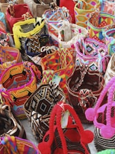 A collection of colorful woven bags with intricate patterns placed closely together. The bags feature a variety of geometric designs and are made with vibrant threads including reds, pinks, yellows, blues, and blacks. Some bags are adorned with tassels or pom-poms.