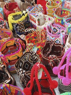 A collection of colorful woven bags with intricate patterns placed closely together. The bags feature a variety of geometric designs and are made with vibrant threads including reds, pinks, yellows, blues, and blacks. Some bags are adorned with tassels or pom-poms.