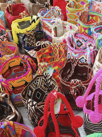 A collection of colorful woven bags with intricate patterns placed closely together. The bags feature a variety of geometric designs and are made with vibrant threads including reds, pinks, yellows, blues, and blacks. Some bags are adorned with tassels or pom-poms.
