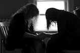 Two people are sitting closely together, with heads bowed and hands clasped, silhouetted against a window. The scene suggests a moment of intimacy or support.