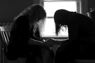 Two people are sitting closely together, with heads bowed and hands clasped, silhouetted against a window. The scene suggests a moment of intimacy or support.