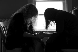 Two people are sitting closely together, with heads bowed and hands clasped, silhouetted against a window. The scene suggests a moment of intimacy or support.