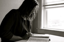 A calm black and white photo of a person reading a book in soft natural light.