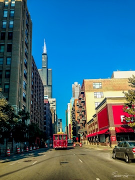 A busy urban street flanked by tall buildings with a red trolley in the center. The Willis Tower is prominently visible against a clear blue sky, and the surrounding structures include brick and glass facades. Cars are parked on the side of the road, and there are orange traffic cones lining the street.