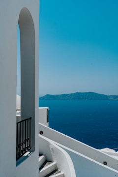A white architectural structure featuring an archway and a staircase with a black railing in the foreground. The background showcases a deep blue sea and a distant, mountainous island under a clear blue sky.