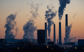 A row of industrial chimneys emit thick plumes of smoke into the sky at sunset, silhouetted against a gradient of orange and blue hues. The scene conveys a sense of industrialization and environmental impact.
