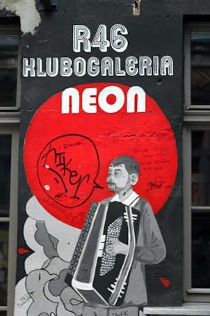 A mural with a monochrome depiction of a person playing an accordion set against a large red circle. The words 'R46 Klubogaleria Neon' are prominently displayed above. The mural includes graffiti-style writing and signatures, adding an urban feel.