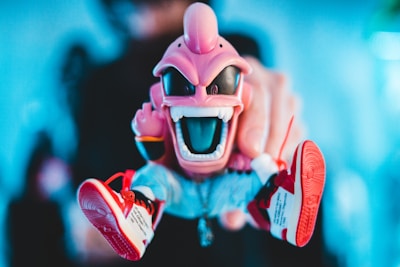 A colorful toy figure with a menacing expression, featuring a wide open mouth and sharp teeth. It is predominantly pink, wearing sunglasses and red sneakers with white soles. A hand is holding the figure in the foreground, set against a soft blue blurred background.