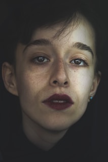 A close-up portrait of a person with short hair and defined eyebrows. The lighting creates a dramatic contrast, highlighting facial features. The expression is serious, with dark lipstick accentuating the lips and small earrings visible.