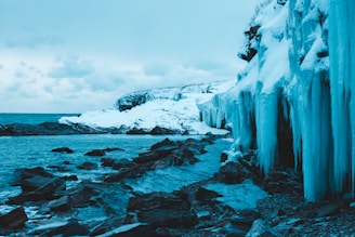 A rugged Greenland coastline with icy waters and a distant mining site under a cloudy sky.