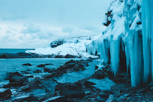 A rugged Greenland coastline with icy waters and a distant mining site under a cloudy sky.