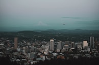 A cityscape with tall buildings in the foreground and a mountain in the distance under a cloudy sky. A drone is flying above the city.