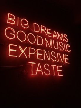 Neon sign displaying the phrases 'BIG DREAMS', 'GOOD MUSIC', 'EXPENSIVE TASTE' illuminated in bright red against a dark background.