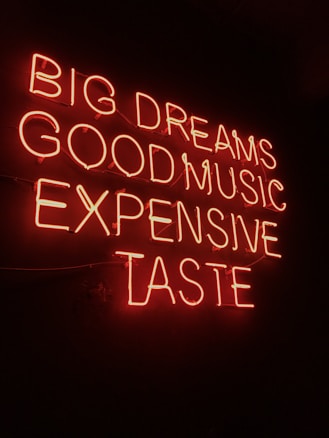 Neon sign displaying the phrases 'BIG DREAMS', 'GOOD MUSIC', 'EXPENSIVE TASTE' illuminated in bright red against a dark background.