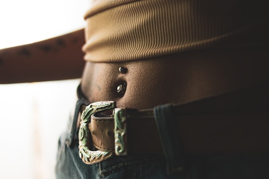 Close-up of a person’s midriff displaying a navel piercing with two metallic balls. The image features a textured belt buckle and denim jeans. The lighting creates a soft, warm tone over the skin and fabric.