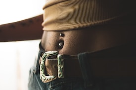 Close-up of a person’s midriff displaying a navel piercing with two metallic balls. The image features a textured belt buckle and denim jeans. The lighting creates a soft, warm tone over the skin and fabric.