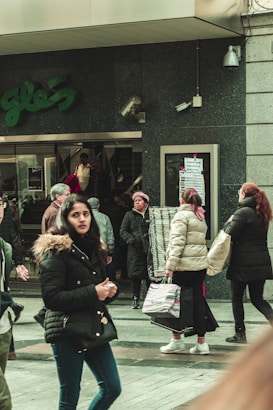 Several people are walking outside a store with a dark facade. One woman in the foreground wears a black coat and looks toward the camera. Other individuals are dressed in winter clothing, some carrying bags. The store entrance has a sign with illegible text, and a mannequin is visible in the window. There is a security camera on the wall.