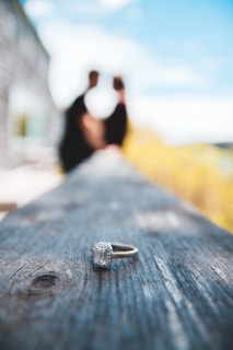 A diamond ring is in sharp focus on a wooden surface, with a blurred background featuring two silhouetted figures facing each other under a bright blue sky.