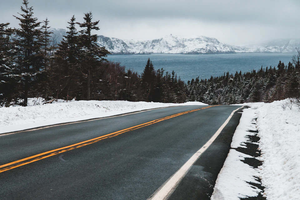 The image depicts a serene winter scene on an empty road, with snow-covered trees lining both sides and a body of water visible in the distance. The sky above is overcast, casting a soft light over the landscape. The perspective of the photo is from the side of the road, looking towards the horizon where the road meets the snowy mountains beyond.