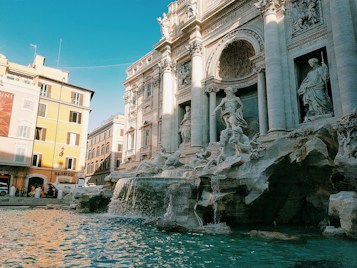 The image features an iconic, intricately designed baroque fountain with grand sculptures. The central figure is a large, impressively carved statue surrounded by smaller figures and stone formations. The water in the foreground reflects the architectural details, creating a serene ambiance. Adjacent to the fountain are historic buildings with classical facades, adding depth to the scene.