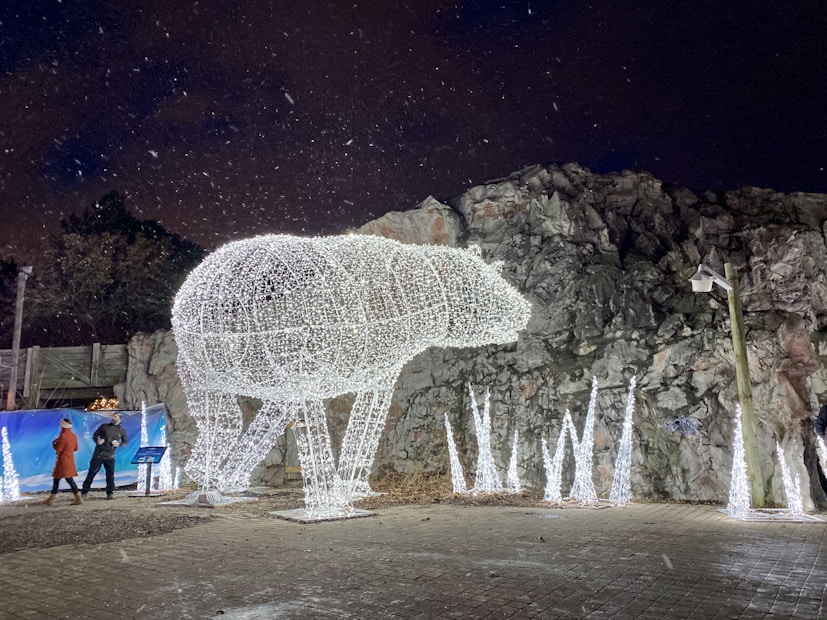 A large illuminated polar bear made of white lights stands against a rocky backdrop. Falling snowflakes add to the ambience, and two people in winter clothing are visible walking nearby. Additional light structures resembling icy stalagmites are positioned around the bear sculpture.