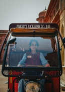 A woman is seated in the driver’s seat of a rickshaw with visible signage on top reading 'Pink City Rickshaw Co. 10'. The background includes traditional architecture with red and tan hues, typical of historic buildings.