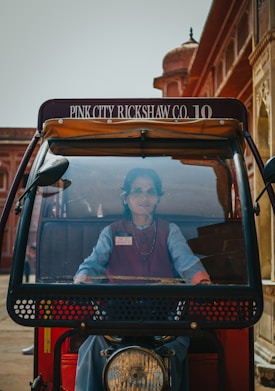 A woman is seated in the driver&rsquo;s seat of a rickshaw with visible signage on top reading 'Pink City Rickshaw Co. 10'. The background includes traditional architecture with red and tan hues, typical of historic buildings.