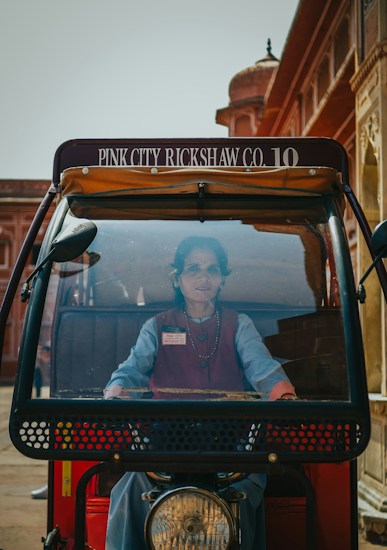 A woman is seated in the driver&rsquo;s seat of a rickshaw with visible signage on top reading 'Pink City Rickshaw Co. 10'. The background includes traditional architecture with red and tan hues, typical of historic buildings.