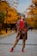 A person stands confidently on a tree-lined path surrounded by vibrant autumn foliage. They are wearing a patterned coat, red sweater, khaki pants, and red shoes. The path is paved with cobblestones and lined with lampposts. The trees display a rich array of orange and yellow leaves, creating a warm, autumnal atmosphere.
