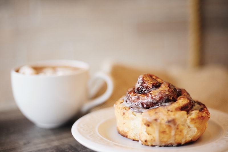 Glazed cinnamon roll on a white plate with a cappuccino cup softly blurred behind it