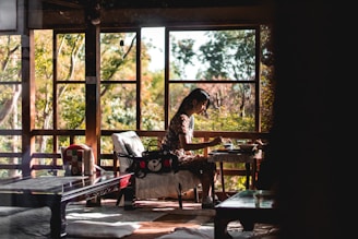 A person is seated at a small table, engaging in an activity that resembles eating or drinking. The setting is a cozy, traditional room with large wooden windows framing lush greenery outside. Soft light filters through the windows, creating a serene and peaceful atmosphere. The decor includes a wooden table with a reflective surface and a cushion on the floor, enhancing the room's rustic charm.