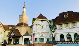 A charming building complex with Bavarian-style architecture, featuring peaked roofs, a clock tower, and decorative façades. The central structure has a sign reading 'Willkommen,' suggesting a welcoming or commercial establishment. Flags are displayed near the entrance, and there are people walking nearby, adding life to the scene.