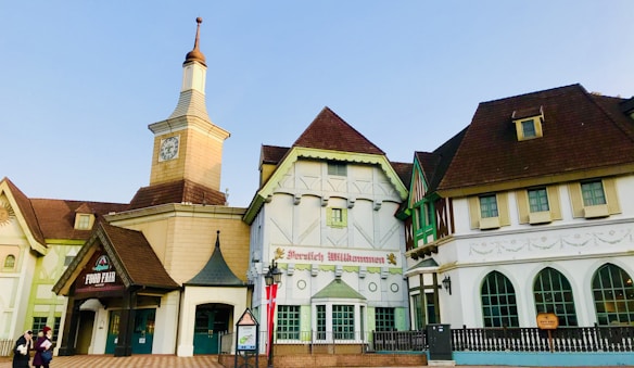 A charming building complex with Bavarian-style architecture, featuring peaked roofs, a clock tower, and decorative façades. The central structure has a sign reading 'Willkommen,' suggesting a welcoming or commercial establishment. Flags are displayed near the entrance, and there are people walking nearby, adding life to the scene.