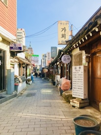 A narrow alleyway lined with traditional Korean buildings and shops featuring various signs in Korean characters. The pavement is made of stone tiles and the street is mostly empty, with a few people visible in the distance. The sky is clear and blue, enhancing the bright and calm atmosphere.