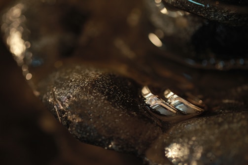 Two silver rings with small diamonds are nestled among smooth, dark rocks that are slightly wet, giving a textured and shiny appearance. The lighting is dim and creates a warm, intimate ambiance with subtle reflections on the metal surfaces.