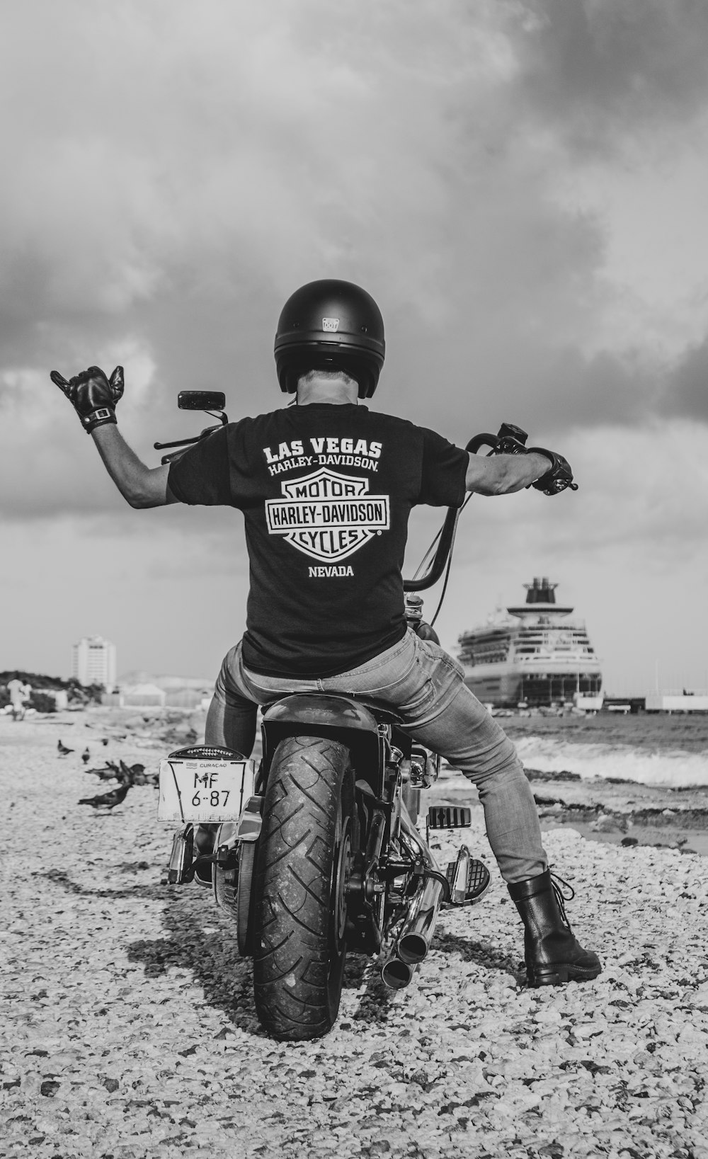 grasyscale photo of man in Harley-Davidson t-shirt riding cruiser motorcycle on shore