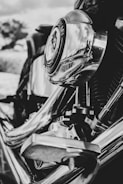 A close-up view of a motorcycle engine featuring shiny chrome parts that reflect the surroundings, including clouds and nature in the background. The intricate mechanical components are clearly visible, with a strong focus on the circular element at the center.