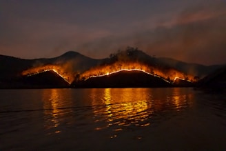 A wildfire rages across a hilly landscape under a dark sky. The flames create a line of bright orange light that contrasts sharply against the dark backdrop. Smoke rises above the hills, and the fiery glow is reflected in the water below, creating a dramatic scene.