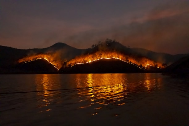 A wildfire rages across a hilly landscape under a dark sky. The flames create a line of bright orange light that contrasts sharply against the dark backdrop. Smoke rises above the hills, and the fiery glow is reflected in the water below, creating a dramatic scene.
