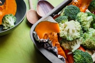 A close-up of a baking tray filled with fresh vegetables including broccoli florets and orange pumpkin slices, with foil wrapped around the pumpkin. A green bowl containing broccoli and wooden spoons can also be seen in the background on a green surface.
