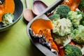 A close-up of a baking tray filled with fresh vegetables including broccoli florets and orange pumpkin slices, with foil wrapped around the pumpkin. A green bowl containing broccoli and wooden spoons can also be seen in the background on a green surface.
