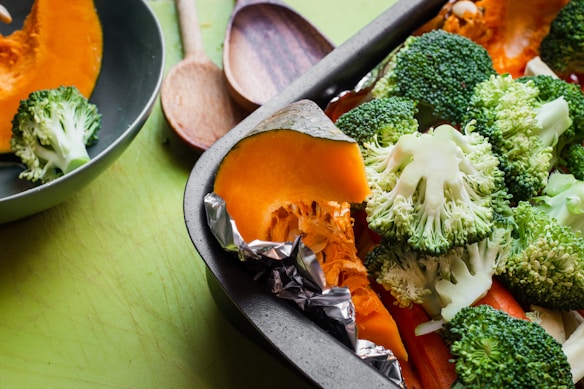 A close-up of a baking tray filled with fresh vegetables including broccoli florets and orange pumpkin slices, with foil wrapped around the pumpkin. A green bowl containing broccoli and wooden spoons can also be seen in the background on a green surface.