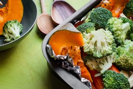 A close-up of a baking tray filled with fresh vegetables including broccoli florets and orange pumpkin slices, with foil wrapped around the pumpkin. A green bowl containing broccoli and wooden spoons can also be seen in the background on a green surface.