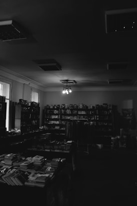 A dimly lit bookstore interior with shelves filled with books against the walls. Tables are arranged in the center, also stacked with books. The room is illuminated by a few hanging light fixtures, casting a moody ambiance.