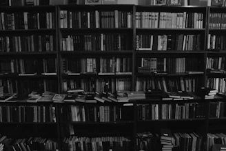 A densely packed bookshelf filled with a variety of books, organized across several shelves and some stacked horizontally. The monochrome tones give the scene a classic and timeless atmosphere, emphasizing the range of book spines, varying in thickness and size.