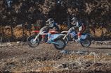 Two motorcyclists are riding off-road bikes over a dirt track, surrounded by a forest setting. Both are wearing helmets and protective gear.