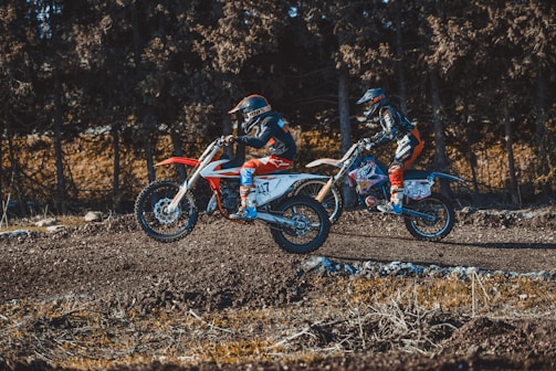 Two motorcyclists are riding off-road bikes over a dirt track, surrounded by a forest setting. Both are wearing helmets and protective gear.