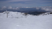 A vast snow-covered landscape features a ski resort with chair lifts ascending a mountain. The scene includes several ski trails extending down the slope, dotted with skiers in the distance. The mountain range is surrounded by a partly cloudy sky, with a mix of sunlight and shadow over the terrain.