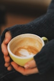 Hands gently holding a green cup of coffee with frothy milk art on top. The person is wearing a cozy dark sweater, suggesting a warm and comfortable scene.