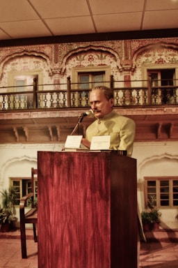 A person standing behind a wooden podium, speaking into a microphone, is dressed in traditional attire. The backdrop features a richly decorated, traditional architectural facade with intricate patterns and a balcony.